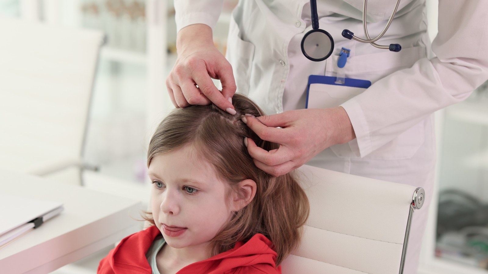 A parent gently checking a young child's scalp for lice by a bright window while a sibling waits nearby