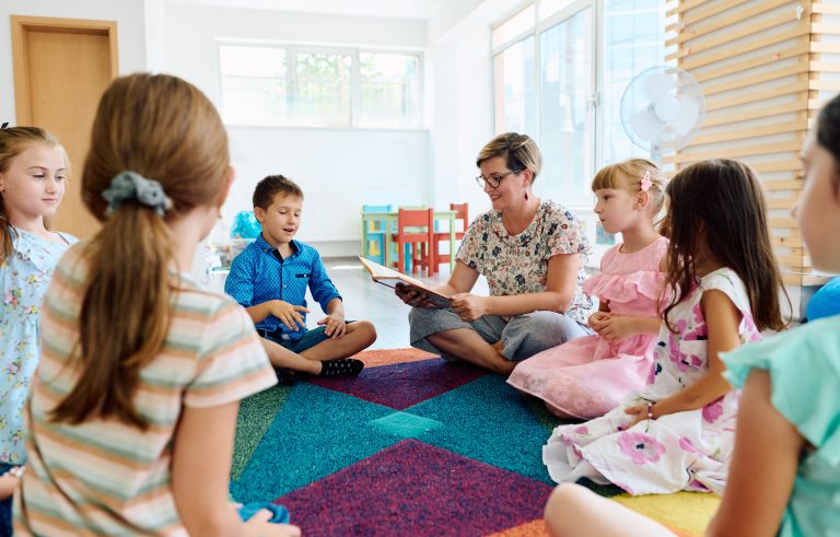 Adult reading a book to children seated on a colorful classroom rug