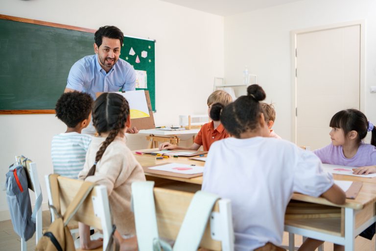 Adult showing a sheet of paper to children seated at classroom tables