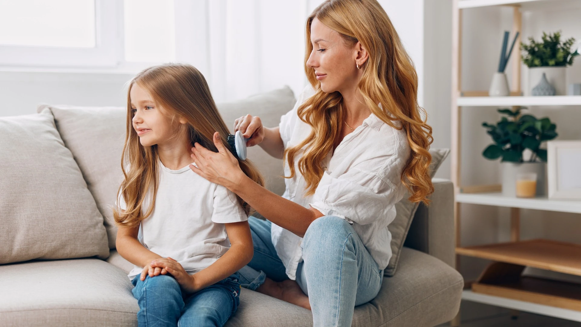 A mother with long wavy blonde hair brushes her young daughter's long honey-blonde hair with a small blue brush as they sit together on a living room sofa